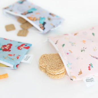 Two snack bags from Bumkins 3-pack (Princess Magic, Ariel, Jasmine) on a white surface: one with heart cookies, the other with crackers.