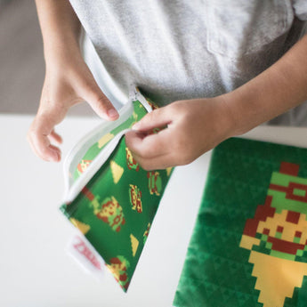 Child in gray shirt opens a green Bumkins Reusable Snack Bag with Legend of Zelda™ pixelated characters, crafted from waterproof fabric.