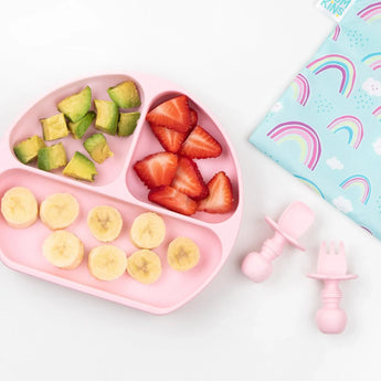 A pink baby plate with fruit sits beside pink utensils, a rainbow cloth, and Bumkins large reusable snack bags: Rainbows & Unicorns.