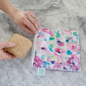 A hand holds a sandwich near a Bumkins Reusable Snack Bag, Large Watercolor pattern, with durable zipper on a marble surface.