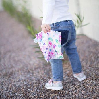 A child walks on gravel with a Bumkins Watercolor & Brush Strokes snack bag, wearing jeans, a white shirt, and sneakers.