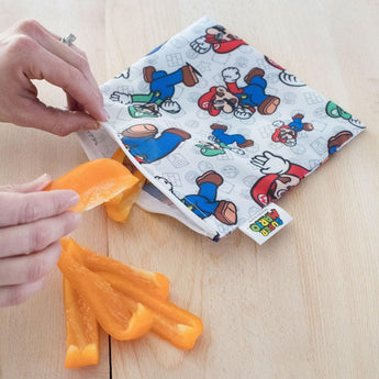 Hands place orange pepper slices into a Bumkins Super Mario™ & Luigi reusable snack bag on a wooden surface.