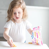 A young girl grabs snacks from a colorful Bumkins Reusable Snack Bag, Watercolor & Brush Strokes design, at a white table.