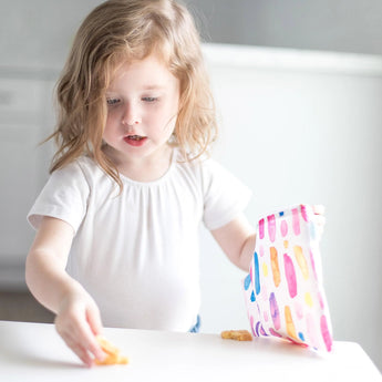 A young girl grabs snacks from a colorful Bumkins Reusable Snack Bag, Watercolor & Brush Strokes design, at a white table.