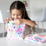 A young girl smiles, holding a Bumkins Reusable Snack Bag, with another vibrant bag from the Large 2-Pack beside her.