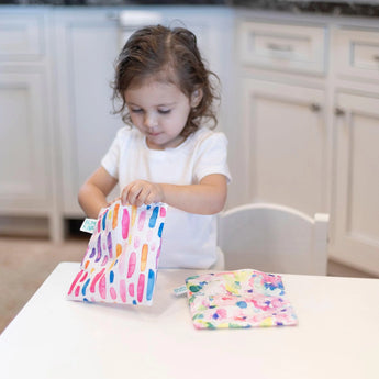 A child joyfully opens a Bumkins Reusable Snack Bag, Large 2-Pack in Watercolor & Brush Strokes, with another vibrant bag by their side.