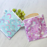 Two Bumkins Reusable Snack Bags on marble: one with rainbows and grapes, the other with unicorns peeking at a sandwich.