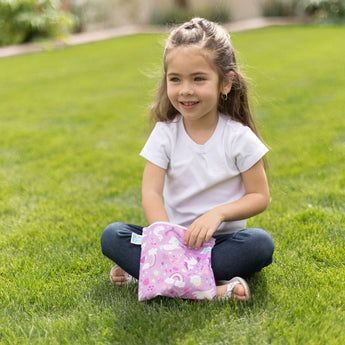 Girl with long hair, wearing a white shirt and jeans, smiles while holding her pink Bumkins Reusable Snack Bag in Rainbows & Unicorns print.