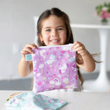 A girl smiles holding a Bumkins Rainbows & Unicorns snack bag made of washable, waterproof fabric; another rainbow bag is beside her.