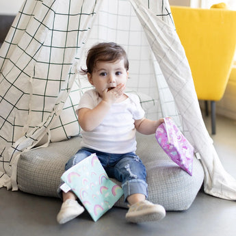 A toddler sits in a fabric tent, holding Bumkins Reusable Snack Bag, Large 2-Pack: Rainbows & Unicorns, and covers their mouth.