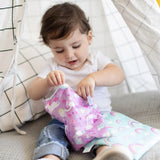 A toddler in jeans plays with a pink Bumkins reusable snack bag featuring rainbow unicorns while sitting inside a tent.