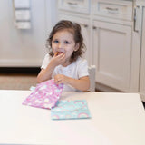 A young girl in a white shirt enjoys snacks from a pink bag at the table with Bumkins Reusable Snack Bag, Large 2-Pack: Rainbows & Unicorns.