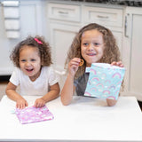 Two smiling children hold Bumkins rainbow-designed Reusable Snack Bags, Large 2-Pack in a kitchen setting.