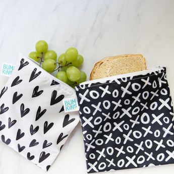 On a marble surface, two Bumkins Reusable Snack Bags from the Large 2-Pack featuring XOXO & Hearts rest; one holds green grapes with a black heart pattern, and the other contains two slices of bread. Made from waterproof fabric, both have durable zipper closures and display BUMKINS tags.