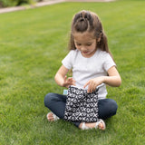 Young girl sitting on grass, wearing a white shirt and holding Bumkins Reusable Snack Bag in XOXO pattern.