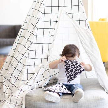 A child in a checkered play tent holds a cloth with XOXO pattern, wearing jeans and white shoes, next to Bumkins snack bags.