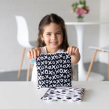 A girl smiles with a black and white XO patterned reusable snack bag from Bumkins, as another sits beside her at the table.