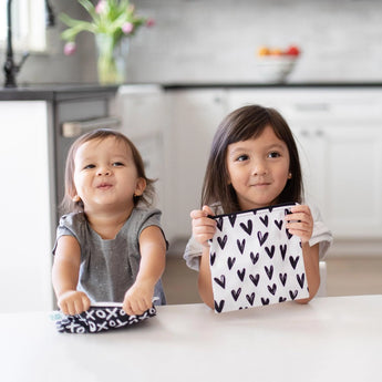 Two kids sit at a white counter, each holding Bumkins Reusable Snack Bags in XOXO & Hearts pattern.