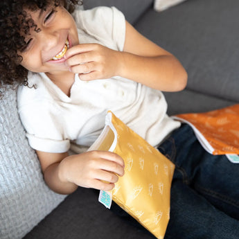 A child in a white shirt and jeans laughs on a couch, holding a Bumkins Reusable Snack Bag from the Sunshine and Grounded 2-Pack.