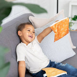 Child smiling, holding a Bumkins Reusable Snack Bag, Sunshine pattern. Another bag from the Grounded set lies on the gray cushion.