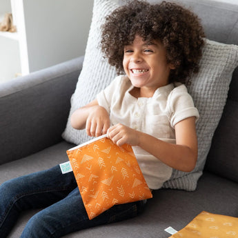 A curly-haired child smiles on a couch, holding a Bumkins Reusable Snack Bag from the Large 2-Pack: Sunshine and Grounded.