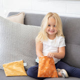 A young girl with long blonde hair and a white shirt sits on a gray sofa, smiling as she holds an orange-patterned pouch from the Bumkins Reusable Snack Bag, Large 2-Pack: Sunshine and Grounded. Another snack bag is beside her, against a large textured gray pillow.