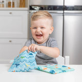 A child smiles at the table, playing with Bumkins ocean life & whale tail reusable snack bags featuring a durable zipper closure.