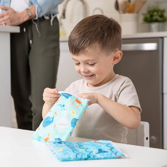 A boy opens a Bumkins Ocean Life snack bag at the table while an adult in the background smiles.