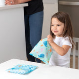 A girl holds a colorful Bumkins Reusable Snack Bag, Large 2-Pack: Ocean Life & Whale Tail; another sits nearby.