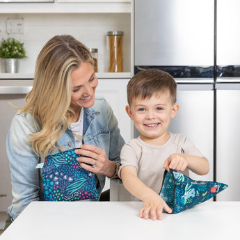 A woman and child sit at a table, smiling with Bumkins Jungle & Animal Prints Reusable Snack Bags made from waterproof fabric.