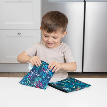 A young boy smiles as he opens a Bumkins Jungle print reusable snack bag with a durable zipper at the kitchen counter.