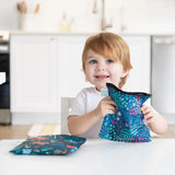 A light brown-haired toddler smiles while holding a Bumkins reusable snack bag with jungle and animal prints at a white table.