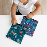 Child in white shirt holding Bumkins Reusable Snack Bag 2-Pack, featuring Jungle & Animal Prints and a sturdy zipper, at a table.