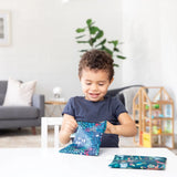 A child beams unwrapping gifts at a table with toys, a plant, and Bumkins large 2-pack reusable snack bags in jungle & animal prints.