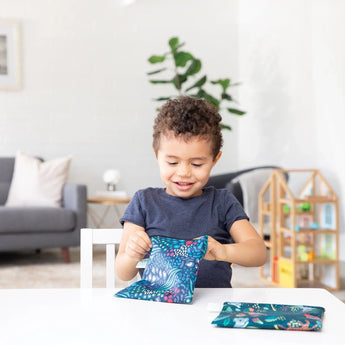 A child beams unwrapping gifts at a table with toys, a plant, and Bumkins large 2-pack reusable snack bags in jungle & animal prints.