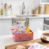 A kitchen counter has a Bumkins 2-pack of Reusable Snack Bags (Watercolor & Brush Strokes), a banana, an orange, and sliced bread.