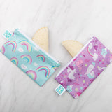 Two Bumkins Reusable Snack Bags from the Rainbows & Unicorns 2-Pack rest on a marble surface. One is teal with rainbows, and the other is purple with unicorns and stars. Made of waterproof fabric, they hold sandwiches with crustless bread corners peeking out.