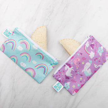 Two Bumkins Reusable Snack Bags from the Rainbows & Unicorns 2-Pack rest on a marble surface. One is teal with rainbows, and the other is purple with unicorns and stars. Made of waterproof fabric, they hold sandwiches with crustless bread corners peeking out.