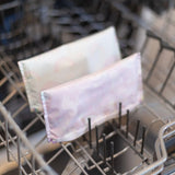 Two pastel-colored Bumkins reusable snack bags on a dishwasher rack, showcasing their machine washable, waterproof fabric design.