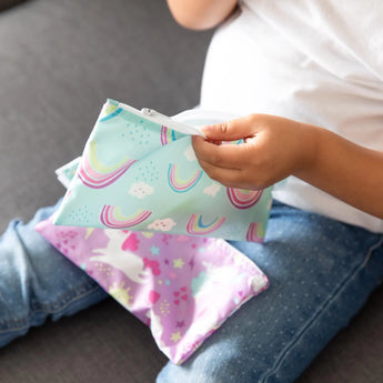 A child holds colorful Bumkins Reusable Snack Bags: Rainbows & Unicorns, both waterproof and machine washable.