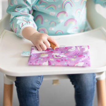 A child in a rainbow outfit has a Bumkins Reusable Snack Bag, Small 2-Pack: Rainbows & Unicorns on their high chair tray.