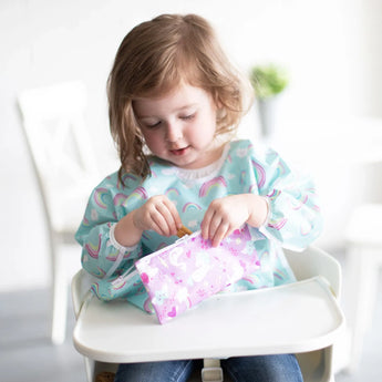 In a bright room, a toddler in a high chair enjoys snacks from Bumkins Reusable Snack Bag featuring rainbows & unicorns.