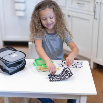 A curly-haired girl wraps her lunch in a reusable wrap. Bumkins small XOXO & Hearts snack bags lie nearby.