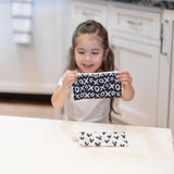 A young girl holds a Bumkins Reusable Snack Bag, while another with black hearts on waterproof fabric lies on the table.