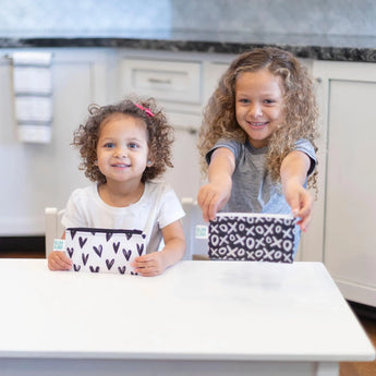 Two kids smile at a table with Bumkins XOXO & Hearts reusable snack bags, perfect for any kitchen.