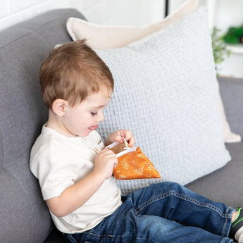 A young boy sits on a gray couch, engrossed in a Bumkins Sunshine-colored Reusable Snack Bag from the Small 2-Pack.