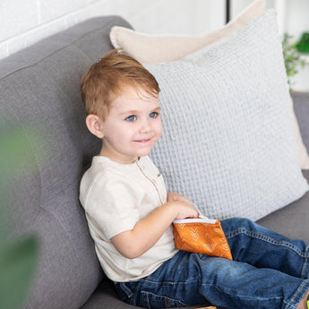 A toddler on a gray sofa smiles, holding a Bumkins waterproof Reusable Snack Bag from the Sunshine and Grounded collection.