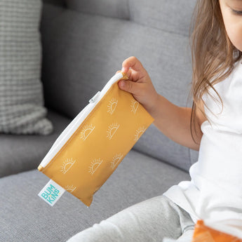 A child on a grey couch holds a small Bumkins Snack Bag with sun designs.