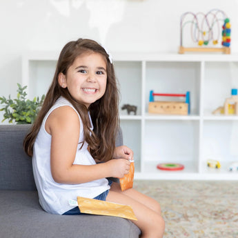 A smiling girl sits on a gray sofa holding a Bumkins Reusable Snack Bag. Shelves with toys are in the background.