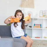 A girl smiles on a gray couch, holding Bumkins Reusable Snack Bags, Sunshine and Grounded 2-Pack. A play area with toys is behind her.